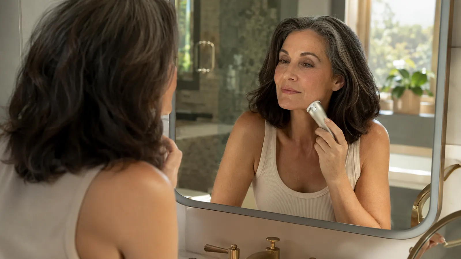 Woman in her mid-fifties applying a microcurrent facial device to her jawline in soft morning light