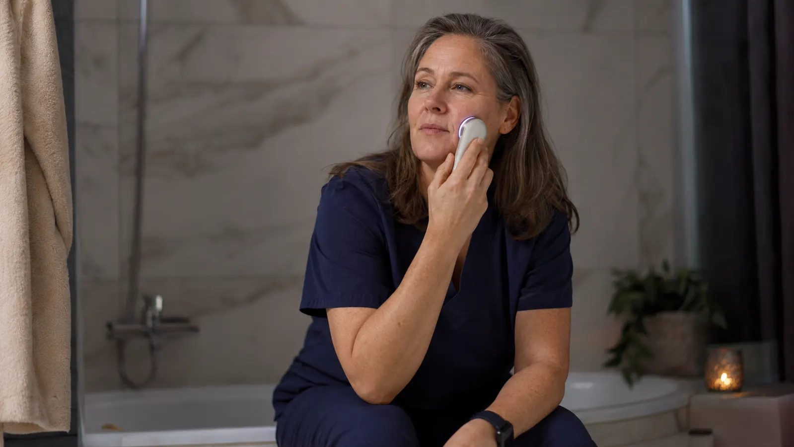 Woman sitting on the edge of a bathtub using a handheld facial device, focused expression