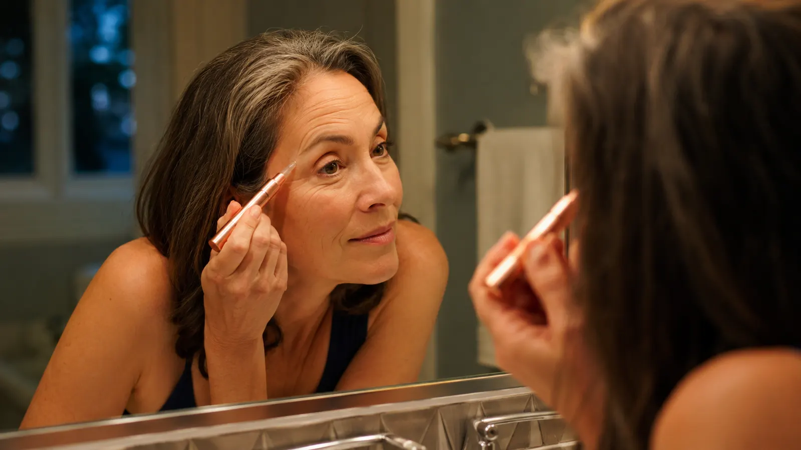 Woman in her fifties leaning toward a bathroom mirror, gently pressing a rose gold red light wand near the corner of her eye