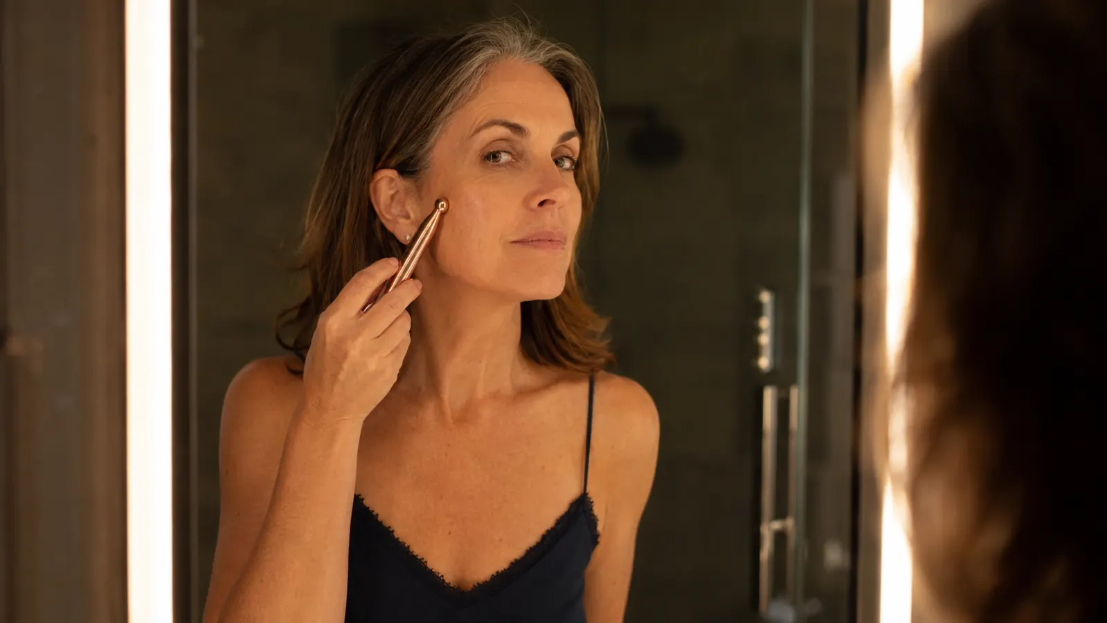 A woman in her fifties gliding a rose gold red light therapy wand along her cheek in warm bathroom light