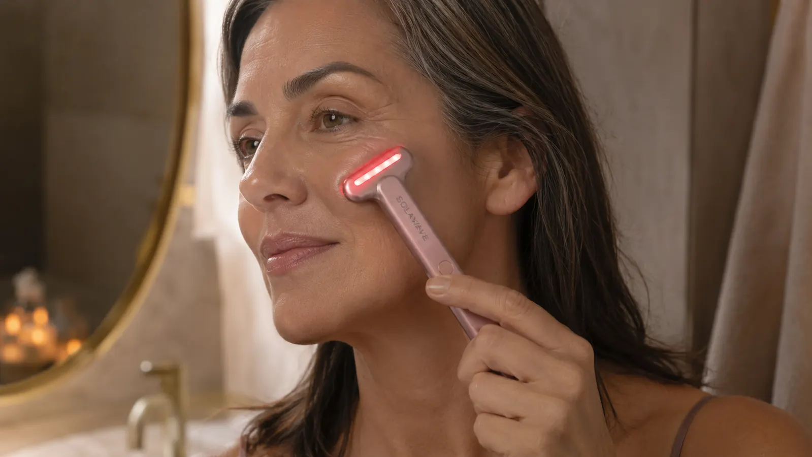 Hand holding the Solawave red light wand against the cheek in front of a bathroom mirror